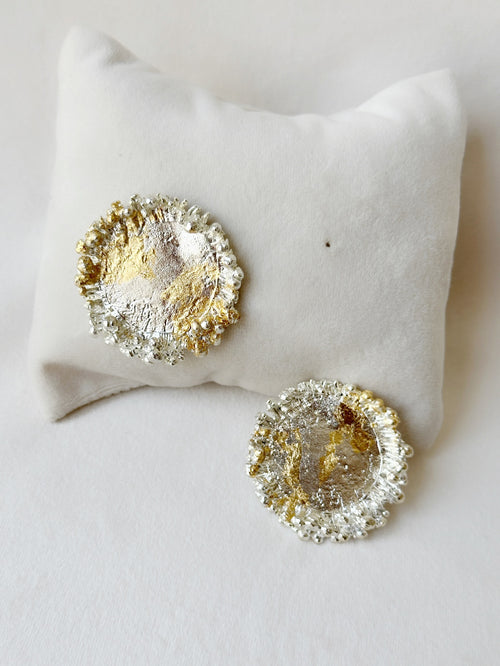 A pair of textured studs earrings, with one half finished in silver and the other in gold, displayed on a white cushion.