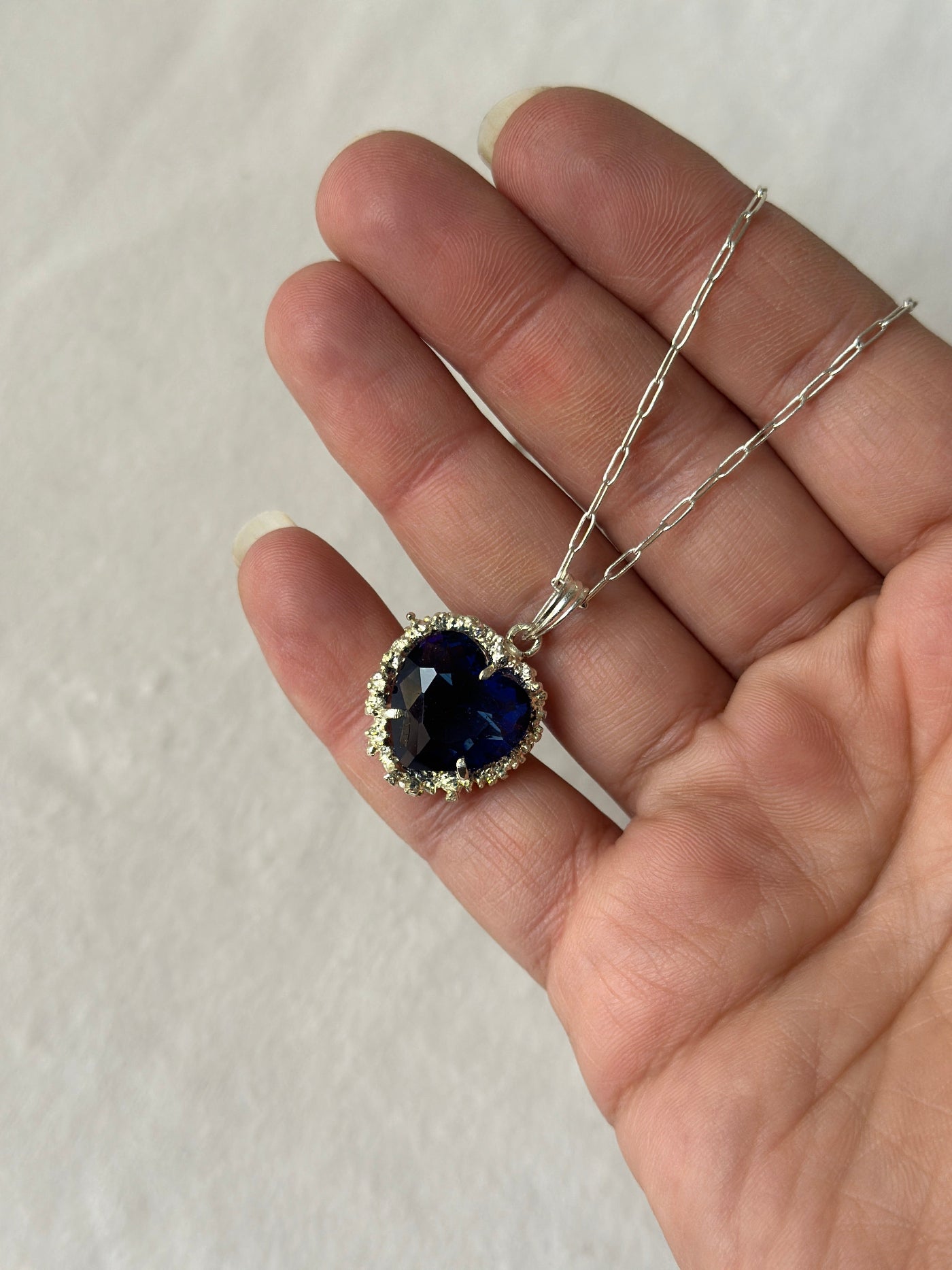 Hand holding a silver necklace with a blue heart-shaped pendant on a light background